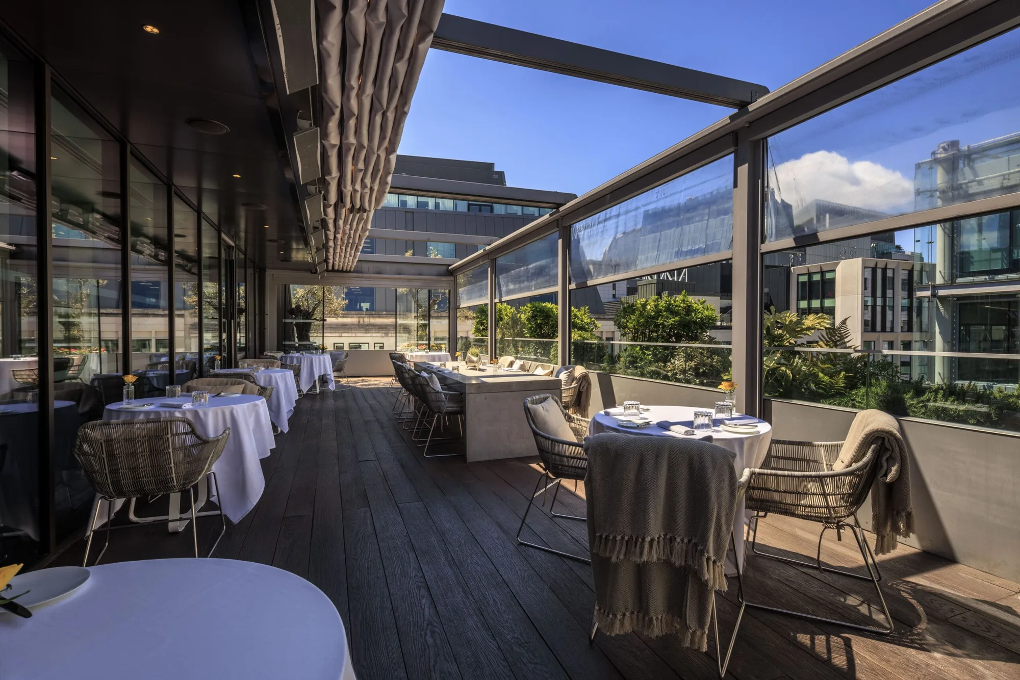 A modern outdoor restaurant terrace with wooden flooring, tables covered in white cloths, and cushioned chairs, some draped with blankets. Glass panels line the sides, and city buildings are visible under a blue sky.