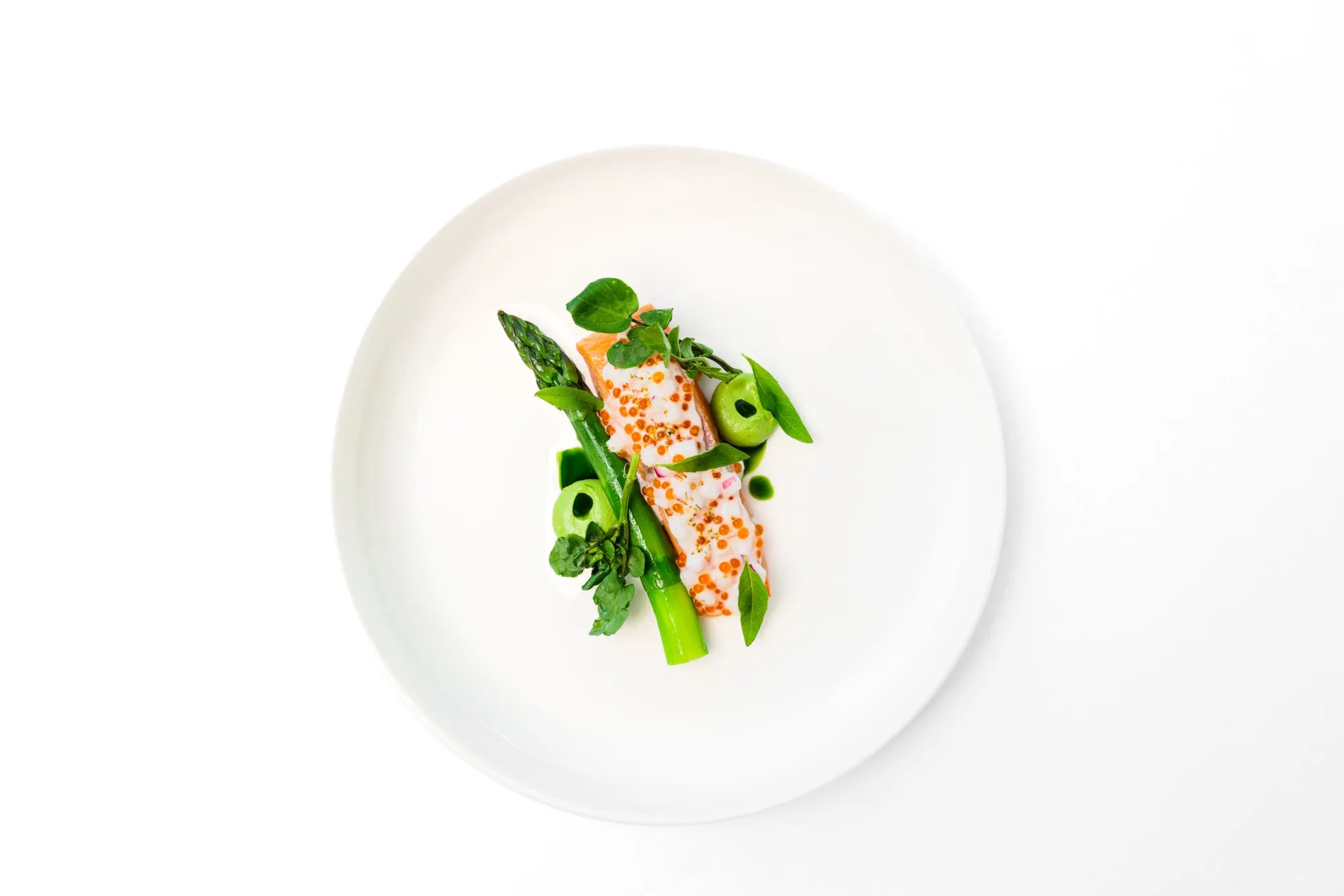 A minimalist dish featuring a piece of fish topped with orange roe, served with asparagus spears, green vegetables, and microgreens on a white plate against a white background.
