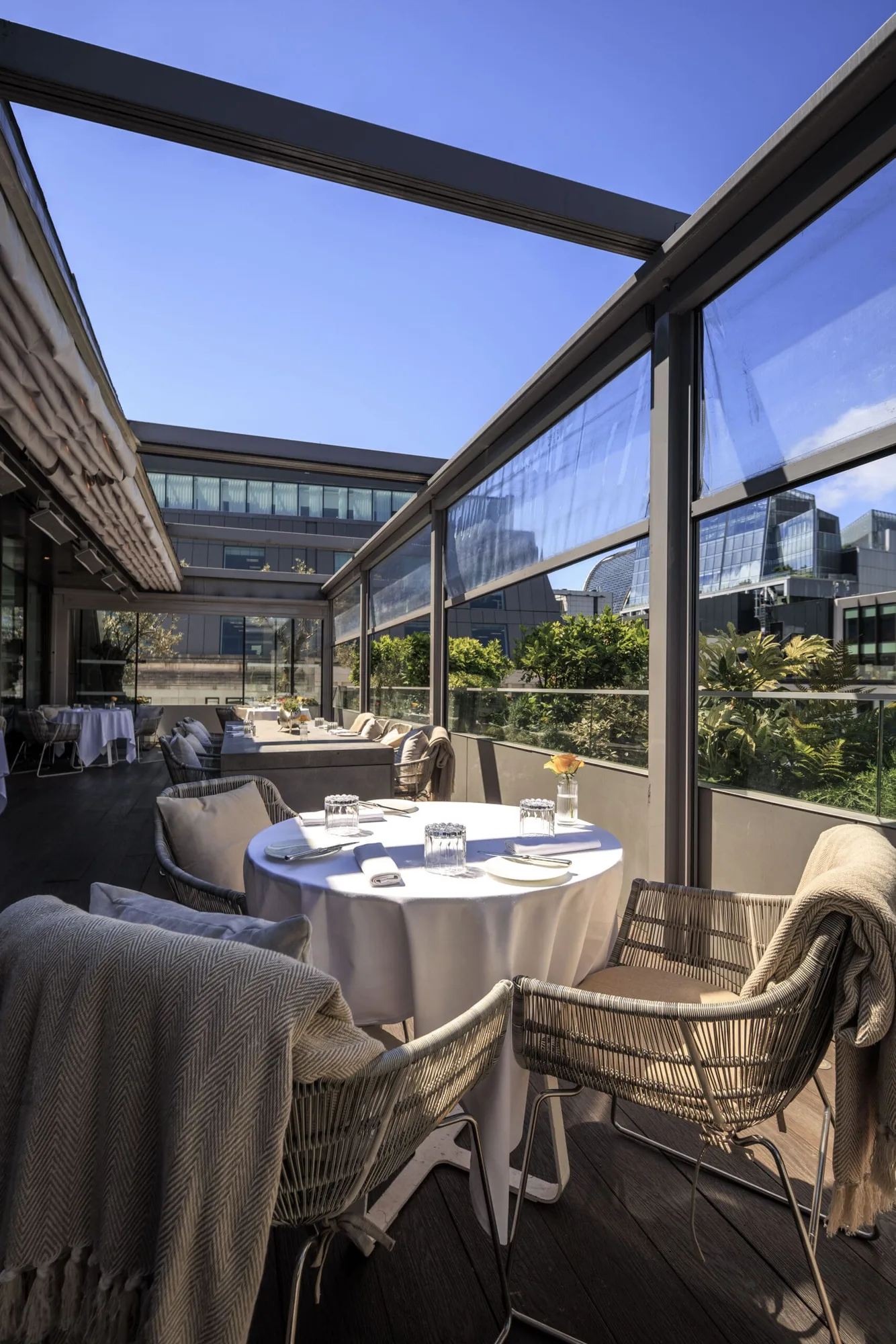Outdoor restaurant terrace with round tables covered in white cloths, wicker chairs, glasses, and plates set for dining. Modern buildings and greenery are visible in the background under a clear blue sky.