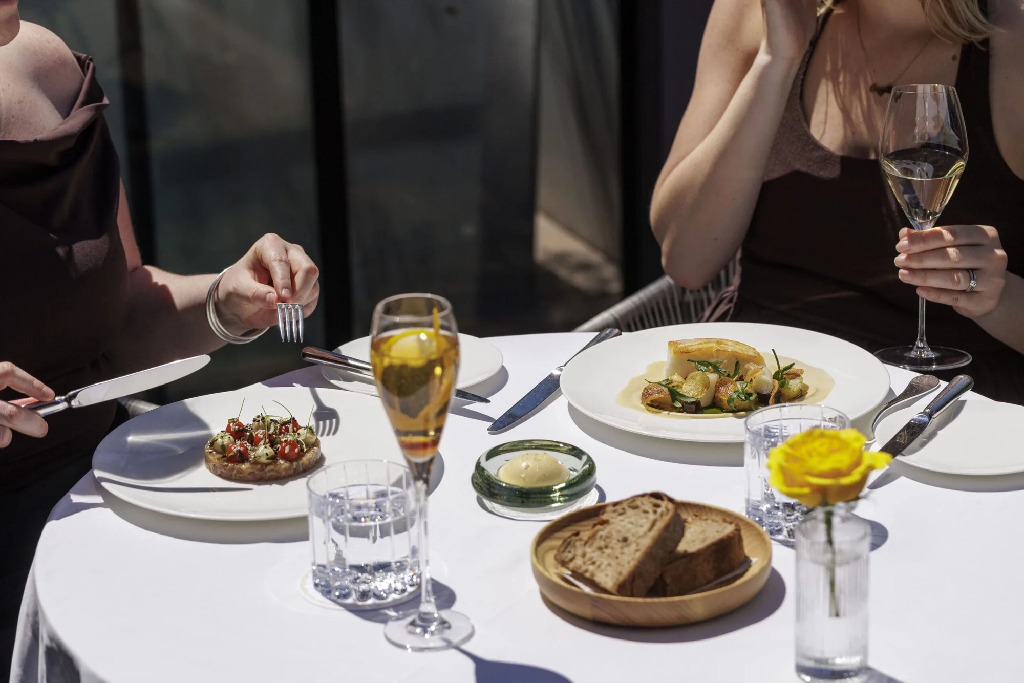 Two women sit at a round, white-clothed outdoor table with plates of gourmet food, glasses of sparkling wine, water, a dish of butter, and a small vase with a yellow flower. Sunlight highlights the elegant setting.