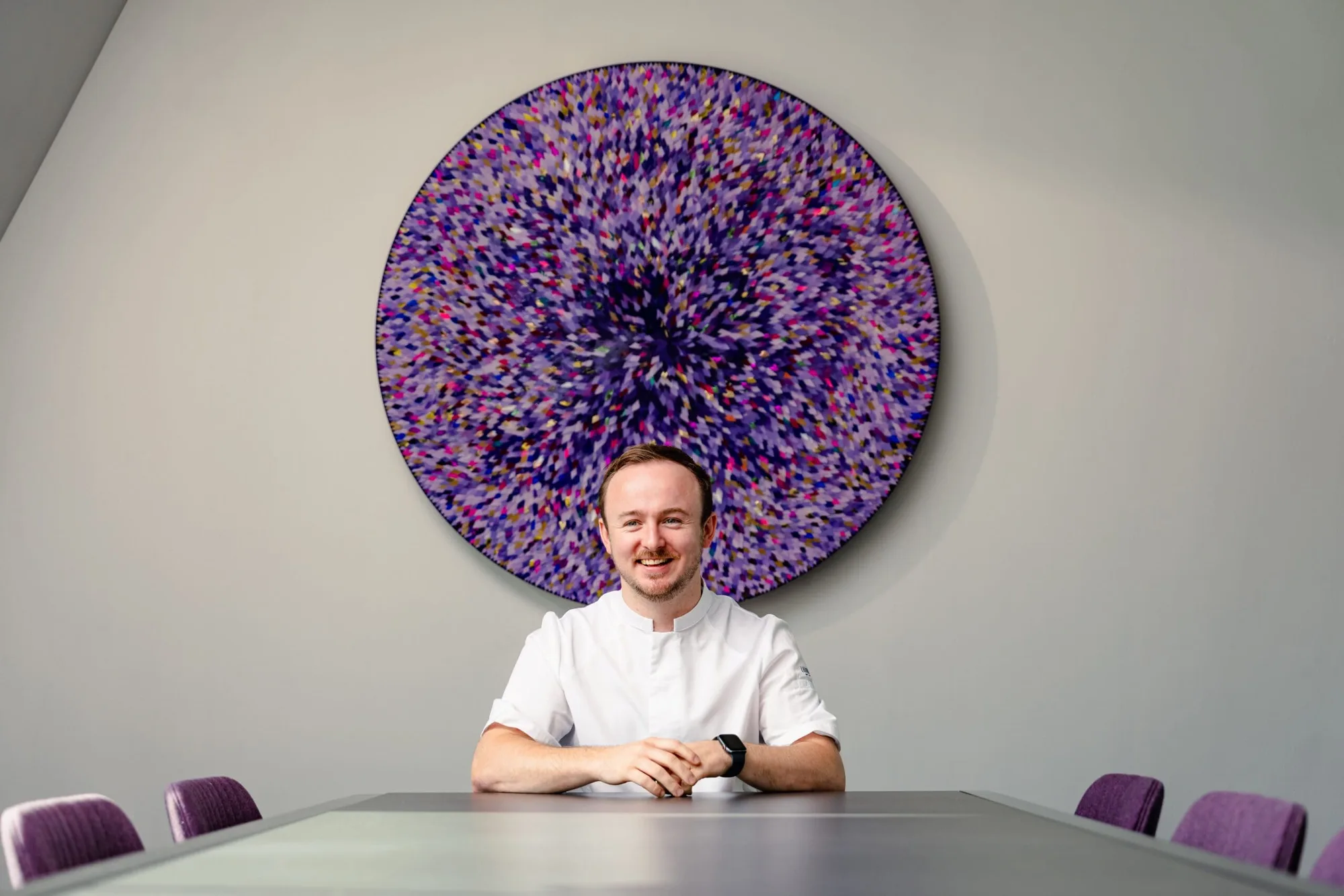 A smiling man in a white shirt sits at a conference table with purple chairs, with a large, circular, abstract artwork in various shades of purple on the wall behind him.