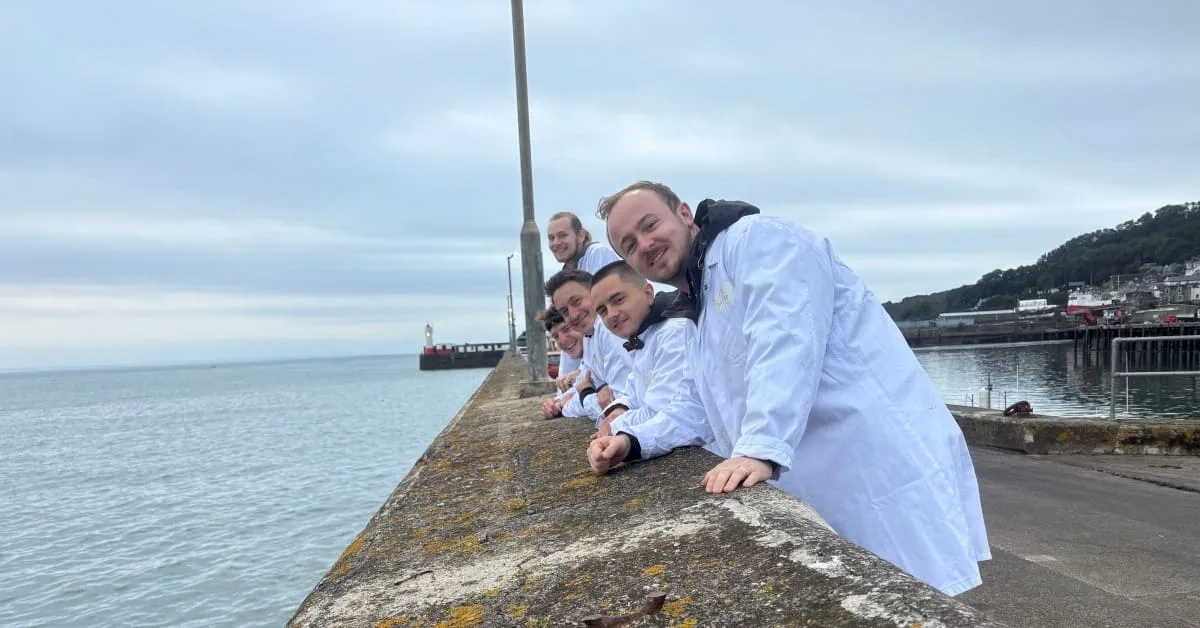 A group of people in white coats lean over a seaside concrete wall, smiling at the camera. The sea and a cloudy sky are in the background, with buildings and a pier visible to the right.