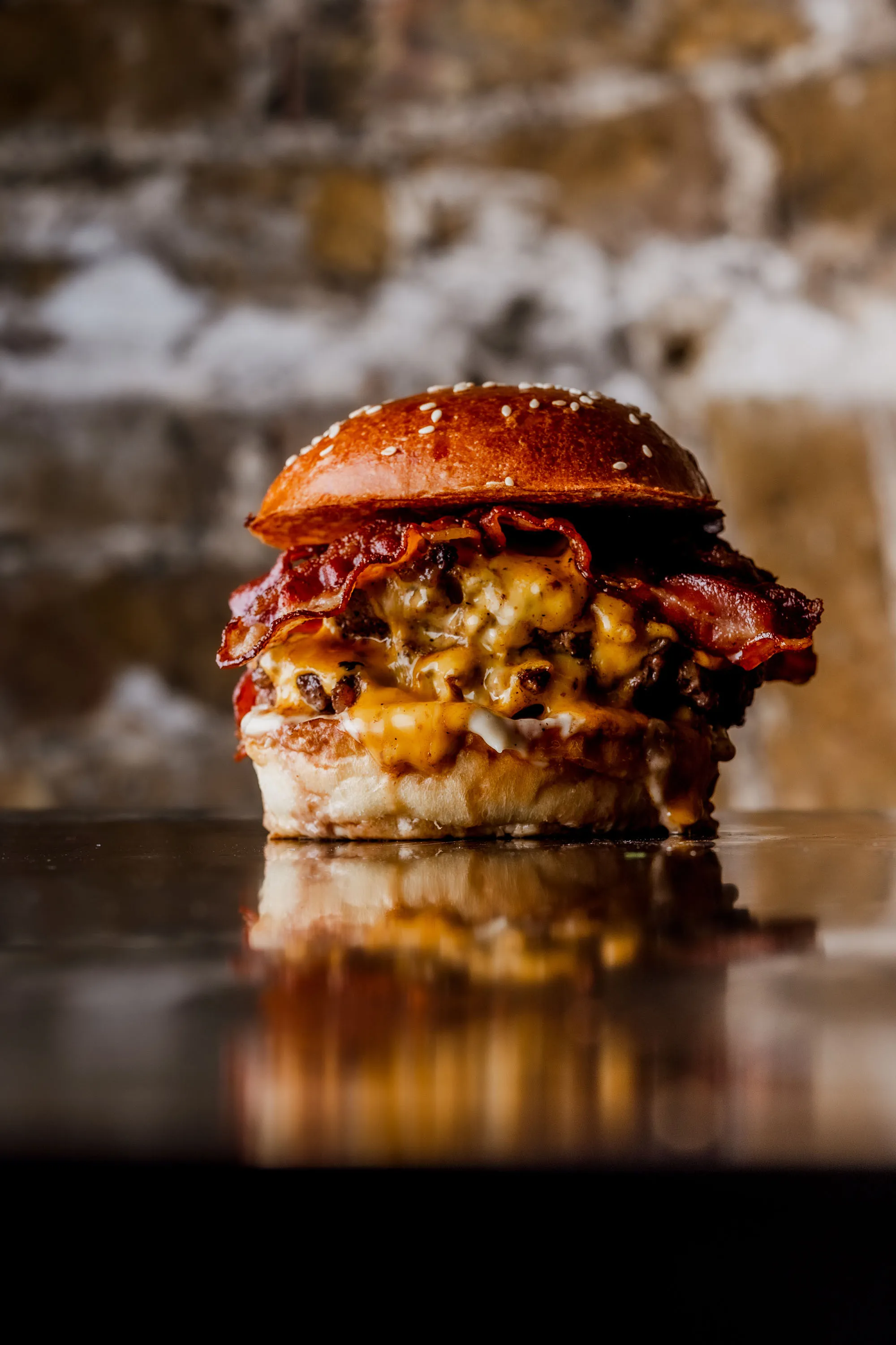 Cheeseburger with bacon and melted cheddar on a sesame seed bun against a blurred stone background.