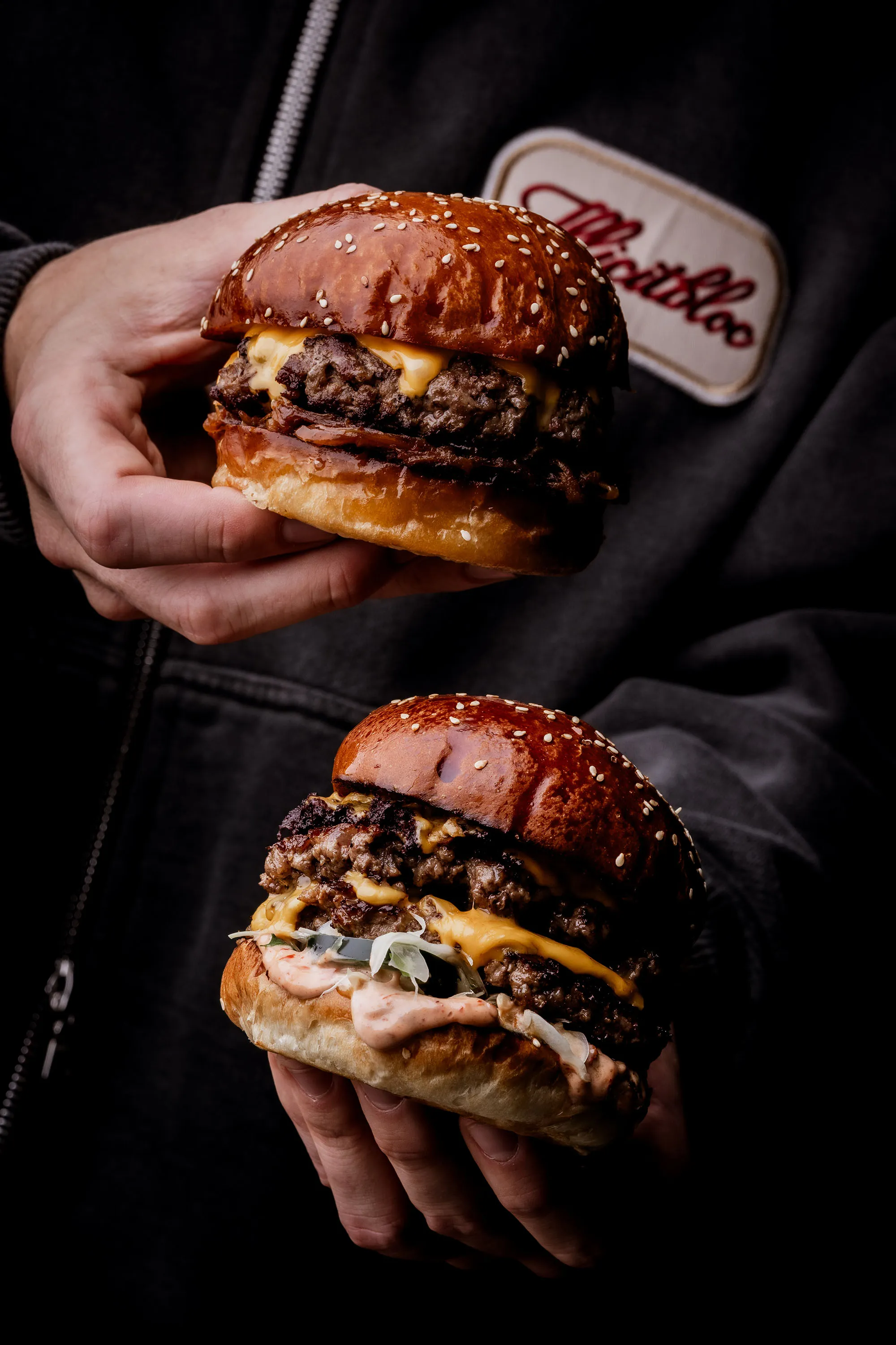 Two hands holding cheeseburgers with sesame seed buns, melted cheese, and toppings against a dark background.