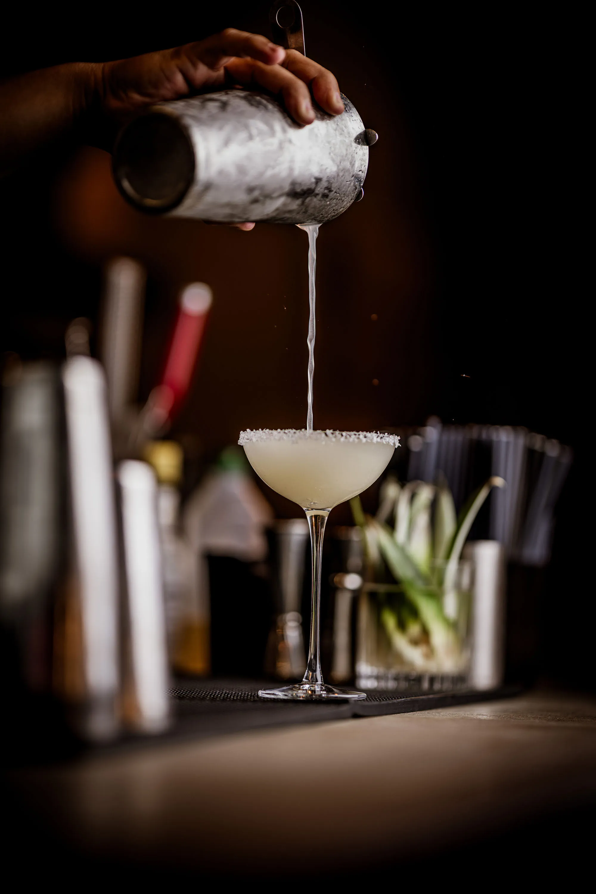 Hand pouring a margarita cocktail from a shaker into a salt-rimmed glass on a bar counter.